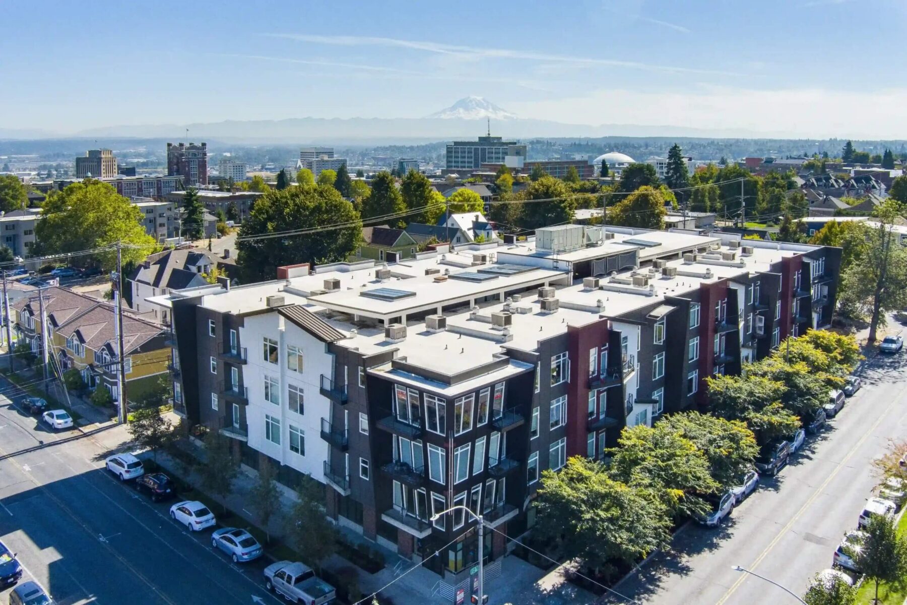 Aerial view of a modern apartment building with white and dark façades, surrounded by trees and parked cars, in an urban neighborhood with a distant mountain visible under a clear sky.