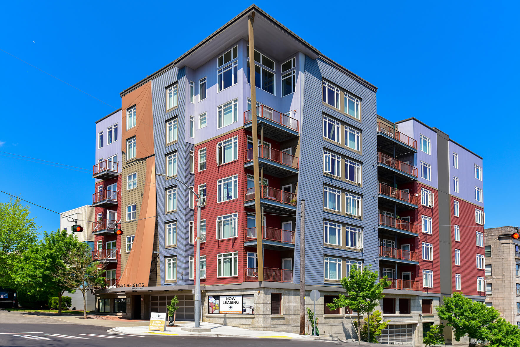 A modern mid-rise apartment building with red and blue exterior panels, large windows, and several balconies, set on a sunny day with a clear blue sky. There are trees and a street in the foreground.